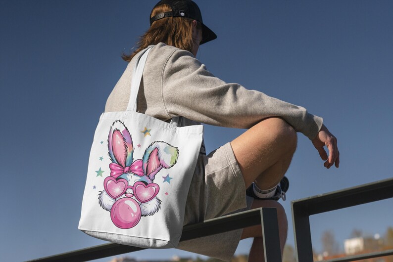 May include: A white tote bag with a colorful cartoon bunny wearing pink heart-shaped sunglasses and blowing a bubble. The bunny has a pink bow on its head and is surrounded by stars.
