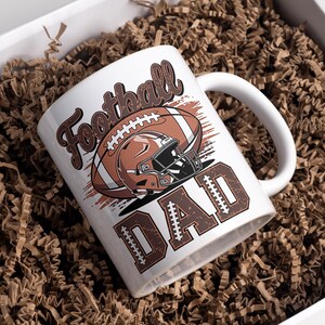 May include: White ceramic mug with a football design. The mug features the words "Football DAD" in brown, along with a football and helmet graphic. The mug is inside a white box filled with brown crinkle paper.