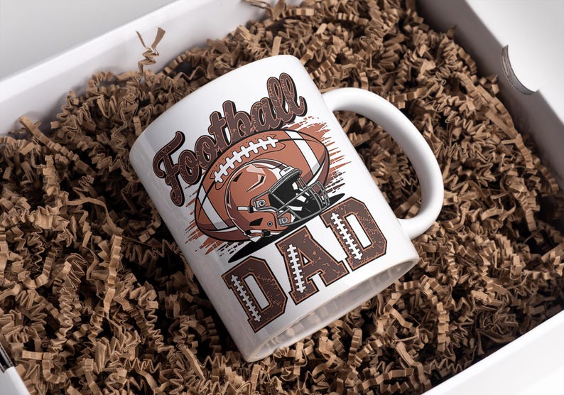 May include: White ceramic mug with a football design. The mug features the words "Football DAD" in brown, along with a football and helmet graphic. The mug is inside a white box filled with brown crinkle paper.