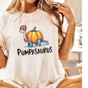 May include: A white t-shirt with a cartoon dinosaur wearing a pumpkin as a hat. The text "Pumpasaurus" is printed below the dinosaur.