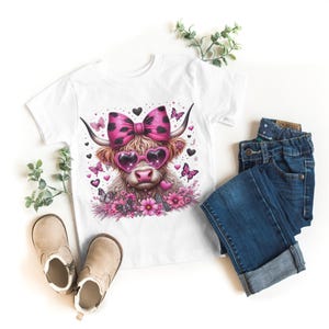 May include: White t-shirt with a pink and black Highland cow wearing pink heart-shaped sunglasses and a pink bow. The cow is surrounded by pink flowers and butterflies.