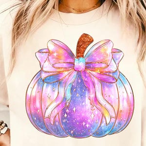 May include: A cream-colored t-shirt featuring a watercolor illustration of a pumpkin with a large, colorful bow. The pumpkin is a mix of purple, blue, and pink with gold accents and sparkles. The bow is a gradient of pink and purple.