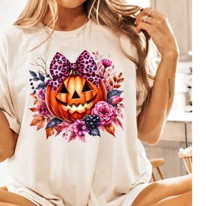 May include: A white t-shirt with a pink and black leopard print bow on top of an orange pumpkin with a jack-o-lantern face. The pumpkin is surrounded by pink, purple, and orange flowers and leaves.