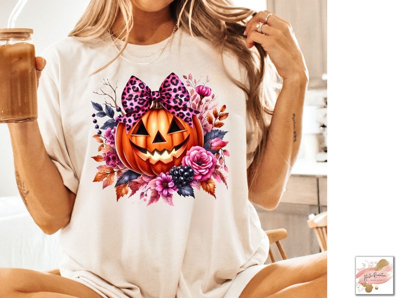 May include: A white t-shirt with a pink and black leopard print bow on top of an orange pumpkin with a jack-o-lantern face. The pumpkin is surrounded by pink, purple, and orange flowers and leaves.