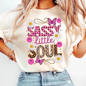 May include: A white t-shirt featuring a pink and black checked graphic that reads "Sassy little soul". The graphic is surrounded by pink and white flowers, butterflies, and smiley faces.