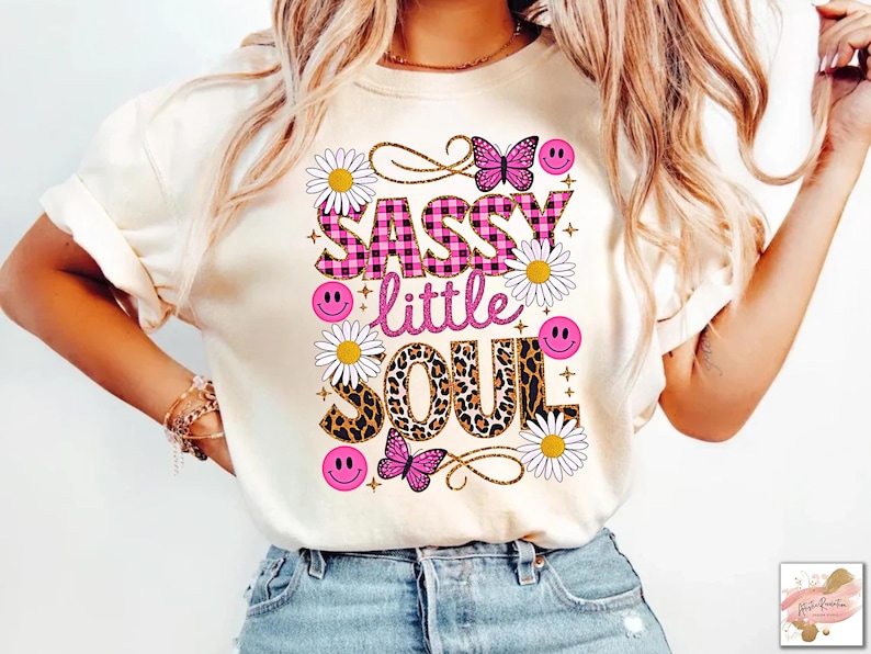 May include: A white t-shirt featuring a pink and black checked graphic that reads "Sassy little soul". The graphic is surrounded by pink and white flowers, butterflies, and smiley faces.