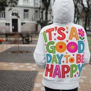 May include: White hoodie with the text "IT'S A GOOD DAY TO BE HAPPY" in colorful, playful lettering. The design includes hearts and a flower. The hoodie is worn outdoors.