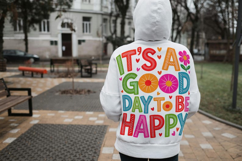 May include: White hoodie with the text "IT'S A GOOD DAY TO BE HAPPY" in colorful, playful lettering. The design includes hearts and a flower. The hoodie is worn outdoors.