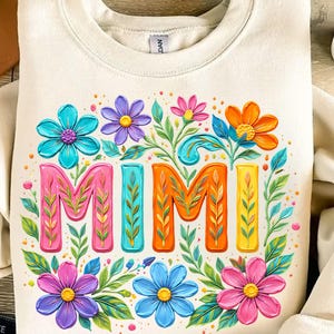 K&ouml;nnte beinhalten: Cremefarbenes Sweatshirt mit einem farbenfrohen Blumendesign und dem Wort "MIMI" in leuchtenden Buchstaben. Das Design zeigt rosa, orange, blaue und lila Blumen mit gr&uuml;nen Bl&auml;ttern, was eine fr&ouml;hliche und auff&auml;llige &Auml;sthetik erzeugt.