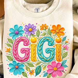 K&ouml;nnte beinhalten: Cremefarbenes Sweatshirt mit dem Wort "GIGI" in farbenfrohen, floralen Buchstaben. Das Design umfasst rosa, t&uuml;rkis, orange und lila Blumen mit gr&uuml;nen Bl&auml;ttern. Das Sweatshirt hat einen Rundhalsausschnitt und lange &Auml;rmel.