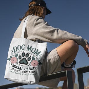 May include: White tote bag with a green and black paw print and floral design. The text on the bag reads "Dog Mom Social Club Life is better with a dog."