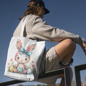 Op de afbeelding: Een witte tote bag met een kleurrijke illustratie van een konijn dat een hartvormig ei vasthoudt. Het konijn wordt omringd door bloemen en andere paaseieren.