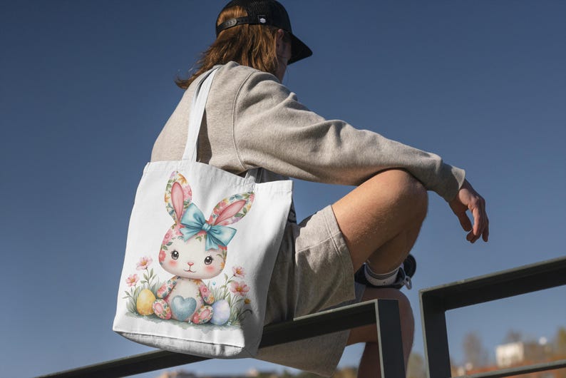 Op de afbeelding: Een witte tote bag met een kleurrijke illustratie van een konijn dat een hartvormig ei vasthoudt. Het konijn wordt omringd door bloemen en andere paaseieren.