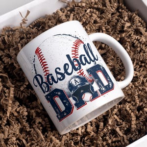 May include: White ceramic mug with a baseball design and the text "Baseball Dad" in blue and red lettering. The mug is sitting in a box filled with brown packing paper.