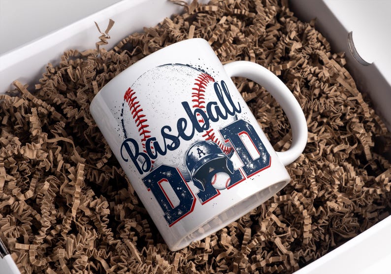 May include: White ceramic mug with a baseball design and the text "Baseball Dad" in blue and red lettering. The mug is sitting in a box filled with brown packing paper.