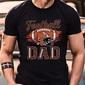 May include: Black t-shirt featuring a football design. The design includes a brown football, a helmet, and the word "Football" in a script font above the football. Below the football is the word "DAD".