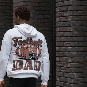 May include: White hoodie with a football-themed graphic on the back. The design features a football, helmet, and the words "Football DAD" in brown and white. The hoodie has a drawstring hood and long sleeves.