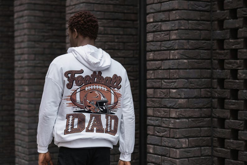 May include: White hoodie with a football-themed graphic on the back. The design features a football, helmet, and the words "Football DAD" in brown and white. The hoodie has a drawstring hood and long sleeves.