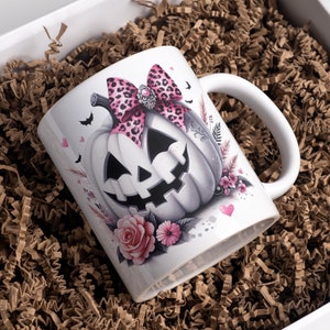 May include: White ceramic mug with a pink and black leopard print bow on a white pumpkin with a spooky face. The mug is surrounded by pink flowers and hearts.