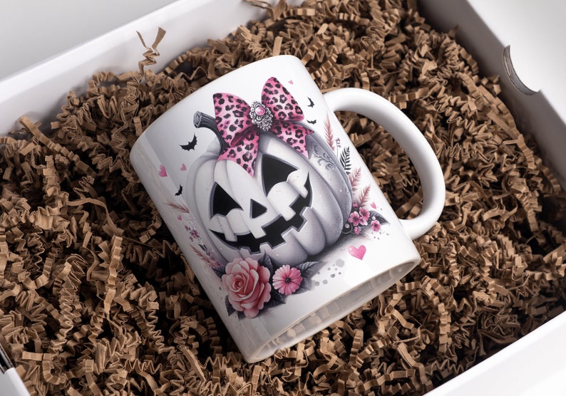 May include: White ceramic mug with a pink and black leopard print bow on a white pumpkin with a spooky face. The mug is surrounded by pink flowers and hearts.