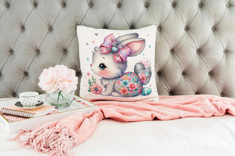 Op de afbeelding: Een wit kussen met een kleurrijke illustratie van een konijn dat een roze strik draagt. Het konijn is omringd door bloemen en heeft een bloemenpatroon op zijn lichaam. Het kussen ligt op een roze deken en een grijs hoofdeinde.