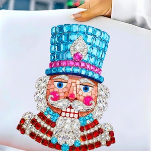 Puede incluir: Un coj&iacute;n decorativo con un dise&ntilde;o detallado de cascanueces enjoyado. El cascanueces tiene un sombrero azul, detalles en rosa y un uniforme rojo y blanco. El dise&ntilde;o est&aacute; hecho de cristales y gemas brillantes, creando un tema festivo navide&ntilde;o.