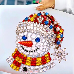 Puede incluir: Un dise&ntilde;o decorativo de mu&ntilde;eco de nieve con sombrero, bufanda y un copo de nieve de colores. El mu&ntilde;eco de nieve est&aacute; adornado con cristales brillantes en varios colores, incluyendo rojo, amarillo, azul y transparente. El dise&ntilde;o est&aacute; sobre un fondo blanco.