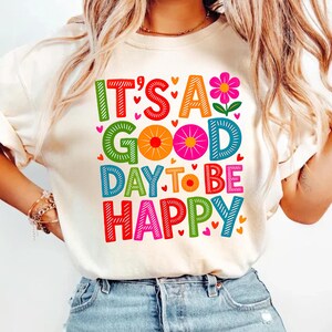 May include: Cream-colored t-shirt with the phrase "IT'S A GOOD DAY TO BE HAPPY" in colorful, block letters. The design includes hearts and a flower. The shirt is being worn by a person.