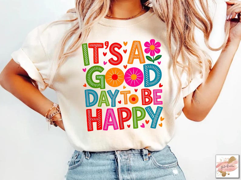 May include: Cream-colored t-shirt with the phrase "IT'S A GOOD DAY TO BE HAPPY" in colorful, block letters. The design includes hearts and a flower. The shirt is being worn by a person.