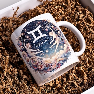 May include: White ceramic mug featuring a Gemini zodiac design. The mug has a handle and is decorated with a celestial illustration of the Gemini constellation, two figures, and the word "Gemini". The mug is presented in a gift box with brown crinkle paper.