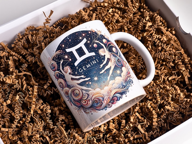 May include: White ceramic mug featuring a Gemini zodiac design. The mug has a handle and is decorated with a celestial illustration of the Gemini constellation, two figures, and the word "Gemini". The mug is presented in a gift box with brown crinkle paper.