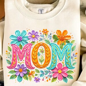 K&ouml;nnte beinhalten: Cremefarbenes Sweatshirt mit einem farbenfrohen Blumendesign und dem Wort &bdquo;MOM&ldquo; in gro&szlig;en, dekorativen Buchstaben. Die Buchstaben sind in Pink, Orange und T&uuml;rkis umrandet, umgeben von Blumen in verschiedenen Farben.