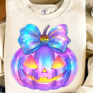 May include: A cream-colored sweatshirt featuring a vibrant, cartoon-style pumpkin design. The pumpkin is purple with a smiling jack-o'-lantern face and a large, blue bow. The bow has a leopard print center and a galaxy-like pattern.