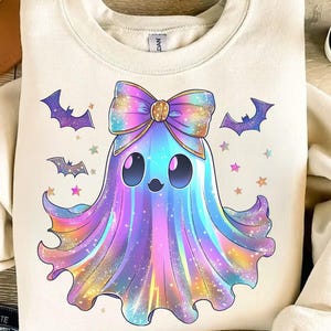 Op de afbeelding: Cr&egrave;mekleurige sweatshirt met een kleurrijk spookontwerp. Het spook heeft een grote strik, vleermuis- en steraccenten en een regenboogkleurig lichaam. Een grillig ontwerp voor Halloween.