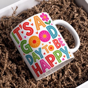 May include: White ceramic mug with the words "IT'S A GOOD DAY TO BE HAPPY" in colorful block letters. The mug features a white handle and is filled with brown packing material inside a white box.