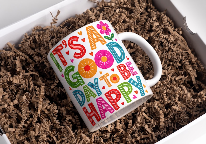 May include: White ceramic mug with the words "IT'S A GOOD DAY TO BE HAPPY" in colorful block letters. The mug features a white handle and is filled with brown packing material inside a white box.