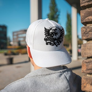 May include: White baseball cap featuring a black and white graphic of an eagle, wings, stars, and the American flag. The cap has a curved brim and is worn by a person in a gray sweatshirt.