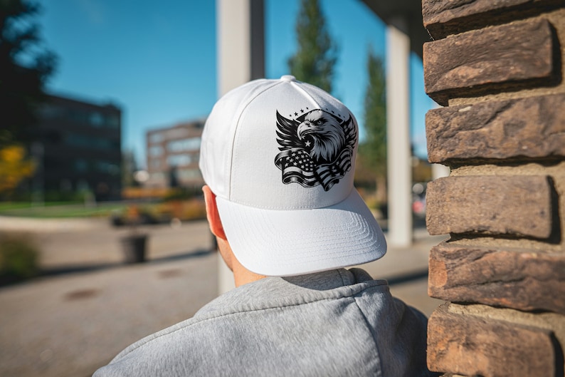 May include: White baseball cap featuring a black and white graphic of an eagle, wings, stars, and the American flag. The cap has a curved brim and is worn by a person in a gray sweatshirt.