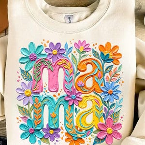 K&ouml;nnte beinhalten: Cremefarbenes Sweatshirt mit dem Wort "Mama" in bunten, floral verzierten Buchstaben. Die Buchstaben sind in Pink, Orange, T&uuml;rkis und Gelb gehalten, umgeben von verschiedenen Blumen in Blau-, Lila- und Rosat&ouml;nen.