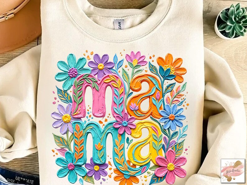 K&ouml;nnte beinhalten: Cremefarbenes Sweatshirt mit dem Wort "Mama" in bunten, floral verzierten Buchstaben. Die Buchstaben sind in Pink, Orange, T&uuml;rkis und Gelb gehalten, umgeben von verschiedenen Blumen in Blau-, Lila- und Rosat&ouml;nen.