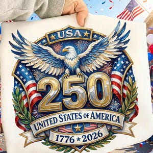 以下が含まれることがあります： 金色のワシが描かれた愛国的なデザインで、「USA 250 UNITED STATES OF AMERICA 1776-2026」の文字が赤、白、青で書かれています。デザインは人が持ち上げており、背景にはアメリカの国旗やお祝いの飾りがあります。