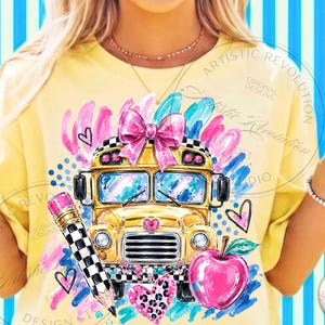 May include: A pale yellow t-shirt featuring a graphic print of a yellow school bus with a large pink bow on top, a black and white checkered pencil, a bright pink apple, and various pink and blue abstract brushstrokes and heart shapes.