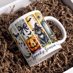 May include: White ceramic mug with a Halloween theme, featuring a black cat in a witch hat, a jack-o'-lantern, and a tombstone. The mug is decorated with bats, a spiderweb, and the words "Spooky Season". The mug is in a white box with brown paper.