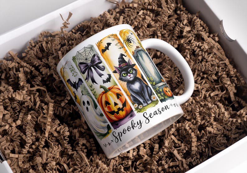 May include: White ceramic mug with a Halloween theme, featuring a black cat in a witch hat, a jack-o'-lantern, and a tombstone. The mug is decorated with bats, a spiderweb, and the words "Spooky Season". The mug is in a white box with brown paper.