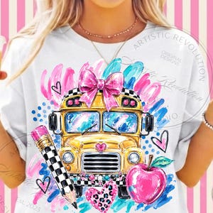 May include: A white t-shirt featuring a graphic print of a yellow school bus with a pink bow on top, a black and white checkered pencil, a red apple, and various pink and blue brushstroke accents and heart shapes on the front.