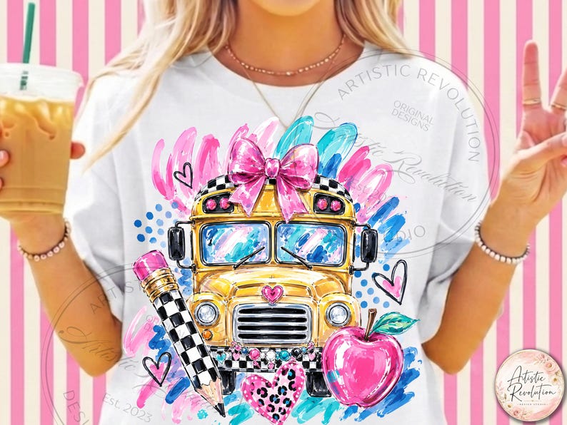May include: A white t-shirt featuring a graphic print of a yellow school bus with a pink bow on top, a black and white checkered pencil, a red apple, and various pink and blue brushstroke accents and heart shapes on the front.