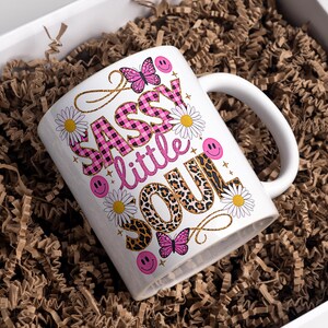 May include: White ceramic mug with a pink, black and gold design. The mug features the text "Sassy little soul" with a pink and black checked pattern, leopard print and daisies.