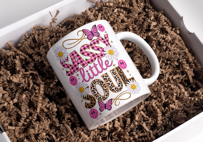 May include: White ceramic mug with a pink, black and gold design. The mug features the text "Sassy little soul" with a pink and black checked pattern, leopard print and daisies.