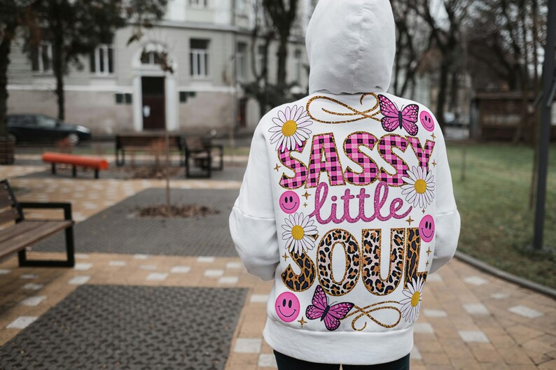 May include: White hoodie with a pink and black graphic print that says "Sassy little soul". The graphic includes flowers, butterflies, and smiley faces.