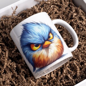 May include: White ceramic mug with a colorful cartoon illustration of a blue bird with an angry expression. The bird has yellow eyes, an orange beak, and orange and brown feathers.